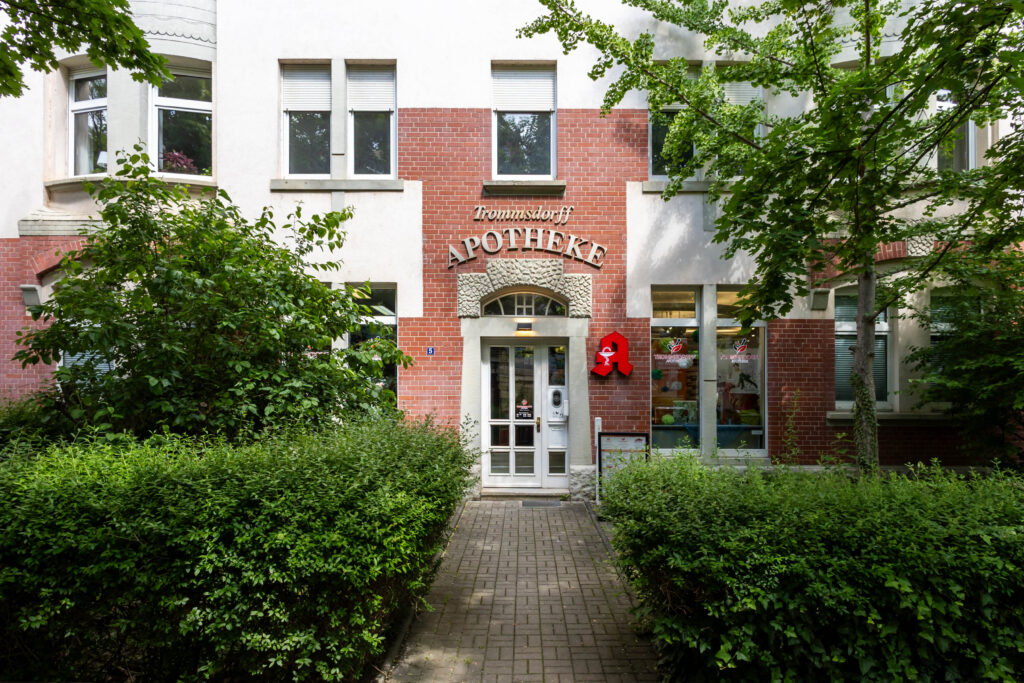 Eingang zur Trommsdorff Apotheke in Erfurt mit einem roten Schild und Pflanzen im Vordergrund. In der Trommsdorff Apotheke in Erfurt